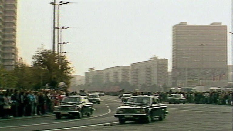 Mehrere Staatskarossen der ehemaligen DDR fahren auf der Straße. Im Hintergrund sind Plattenbauten zu sehen. Am Straßenrand stehen zahlreiche Menschen. (Foto: SWR)