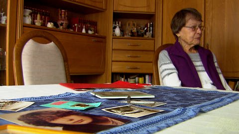 Im Vordergrund Bilder auf einem Tisch, dahinter eine ältere Frau. Der ungeklärte Mordfall Sabine Rahn beschäftigt Angehörige und Ermittler auch nach 40 Jahren noch. (Foto: SWR)