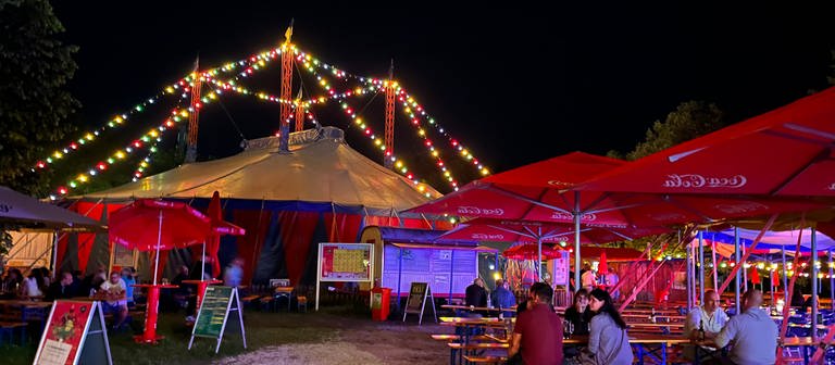 Ein bunt beleuchtes Zirkuszelt, davor Bierbänke mit Publikum. Das Ulmer Zelt im Jahr hat das Programm für die Saison 2023 vorgestellt (Archivbild). (Foto: SWR)
