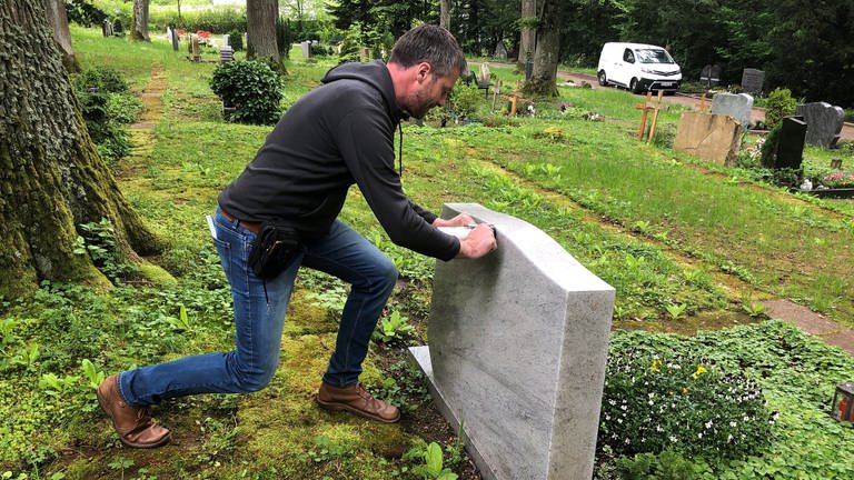 Ein Mann drückt mit Kraft gegen einen hellgrauen Grabstein auf einem Friedhof in Aalen, im Hintergrund Bäume, Gräber und Wiese. (Foto: SWR, Frank Polifke)