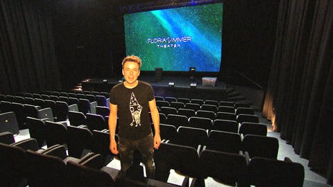 Florian Zimmer steht im Showroom seines Theaters zwischen den leeren Zuschauersesseln. Im Hintergrund ist sein Name auf einem großen Bildschirm zu sehen. Zimmer hofft, nach dem Brand schnell wieder öffnen zu können. (Foto: SWR)