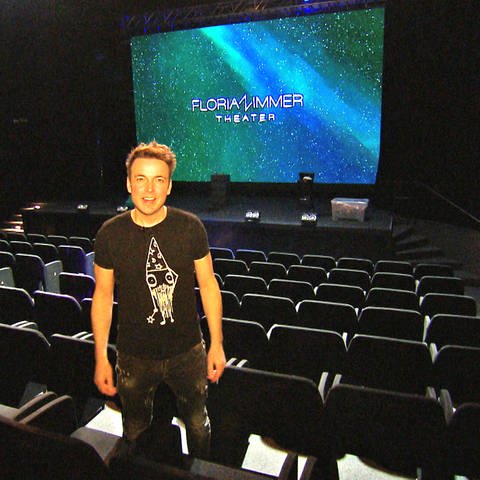 Florian Zimmer steht im Showroom seines Theaters zwischen den leeren Zuschauersesseln. Im Hintergrund ist sein Name auf einem großen Bildschirm zu sehen. Zimmer hofft, nach dem Brand schnell wieder öffnen zu können. (Foto: SWR)