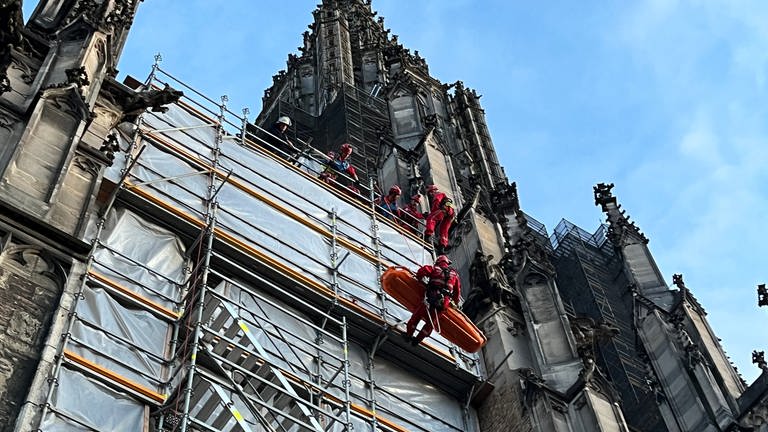 Höhenretter der Feuerwehr seilen an einem Gerüst am Ulmer Münster eine orangefarbene Trage ab. Am Münster in Ulm hat es eine Übung für Höhenrettung gegeben. An einer Trage seilten die Höhenretter eine Trage ab. (Foto: SWR, Markus Bayha)