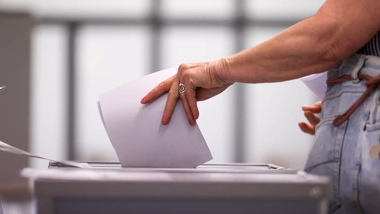 Eine Hand steckt Wahlunterlagen in eine Wahlurne. Am Sonntag werden bei der OB-Wahl 2023 die Ulmerinnern und Ulmer an die Wahlurnen gebeten, um eine neue Oberbürgermeisterin oder einen Oberbürgermeister zu wählen (Symbolbild).  (Foto: dpa Bildfunk, Jan Woitas (Symbolbild))