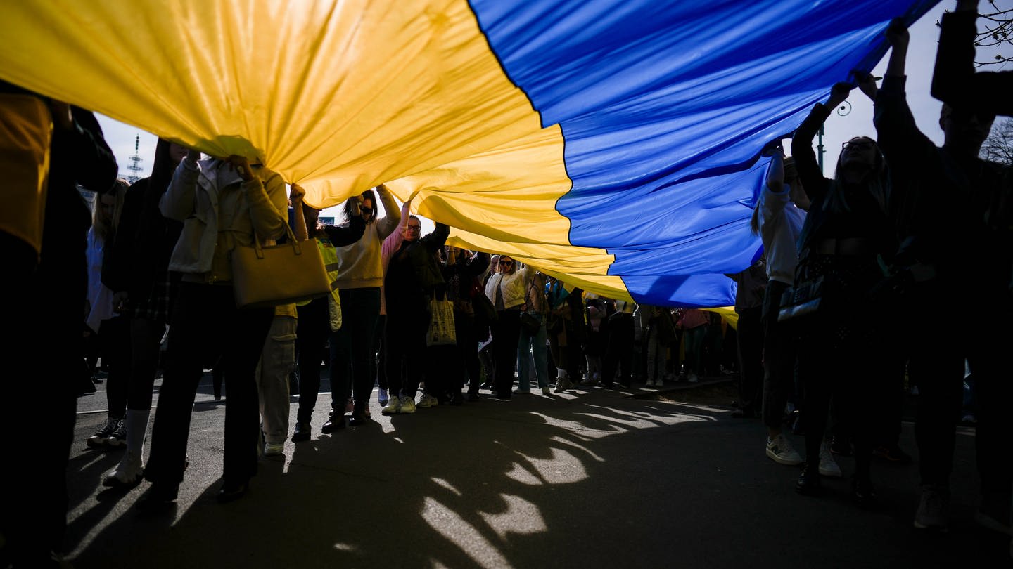 Demonstrierende halten eine große Ukraine-Flagge hoch, das Sonnenlicht scheint von oben hindurch. Der Angriffskrieg auf die Ukraine jährt sich bereits zum zweiten mal. Seitdem werden sowohl Friedensaktivisten als auch Rüstungshersteller in einem andern Licht gesehen. (Foto: picture-alliance / Reportdienste, Picture Alliance)