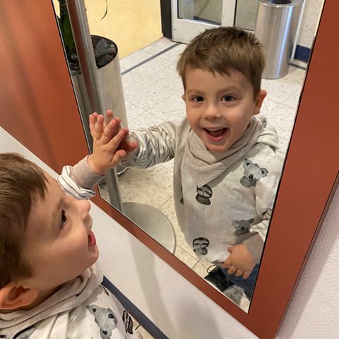 Ein Junge schaut in einen Spiegel und lacht dabei.Theodor (4 Jahre) hat das PURA-Syndrom. Sein Körper produziert ein fehlerhaftes Protein, das in den Zellen Probleme macht.  (Foto: privat)