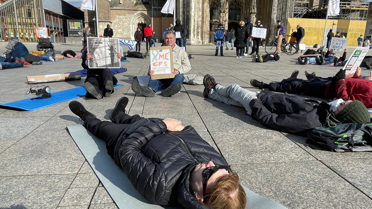 Menschen liegen auf dem Münsterplatz in Ulm, im Hintergrund das Münster. Sie demonstrieren im Liegen, um auf die Krankheit ME/CFS aufmerksam zu machen. Betroffene sind oft zu erschöpft, um das Haus zu verlassen. (Foto: SWR, Janke)