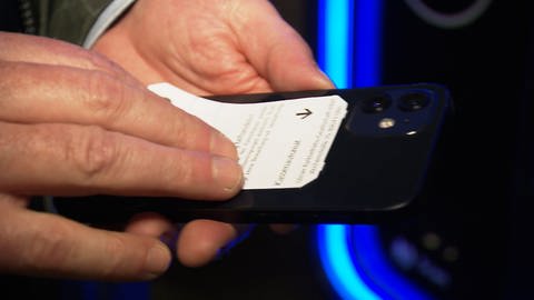Ein Mann hält ein Parkticket an ein Smartphone. Wenn das Parkticket ans Handy kommt, zerstören dessen Magneten die Informationen auf dem braunen Magnetstreifen. Die Ausfahrt aus dem Parkhaus in Ulm ist dann nicht mehr möglich. (Foto: SWR)