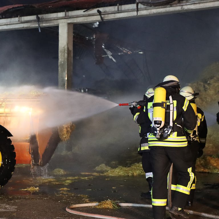 Feuerwehrleute löschen vor einer ausgebrannten Halle einen Traktor. Beim Brand in einer Lagerhalle in Holzheim-Ellerbach wurde unter anderem eine Fotovoltaik-Anlage zerstört.