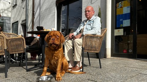 Uwe Schmid sitzt draußen in einem Cafe mit einer Tasse Kaffee. Vor ihm sitzt seine Bordeauxdogge Eddy mit einem Wassernapf. Beide genießen die Gesellschaft im Café. (Foto: SWR, Justus Madaus)