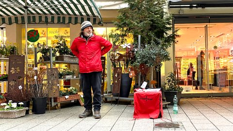 Ein Mann in roter Jacke und Strickmütze steht in einer Gasse vor einem Geschäft. Jörn "Lemmy" Zweng verkauft die Obdachlosenzeitung in der Platzgasse in Ulm. (Foto: SWR, Hannah Schulze)