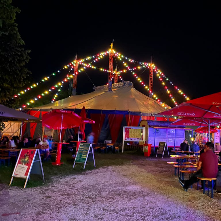 Ein bunt beleuchtes Zirkuszelt, davor Bierbänke mit Publikum. Das Ulmer Zelt im Jahr hat das Programm für die Saison 2023 vorgestellt (Archivbild). (Foto: SWR)
