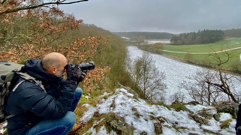 Ein Mann blickt mit einer Kamera in der Hand übers Lonetal. Tierfotograf Jochen Bayer auf dem Hubertusfelsen, wo Wanderfalken brüten.  (Foto: SWR, Anita Schlesak)