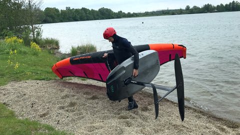 Jürgen Schmid hat einen Neoprenanzug an und läuft mit seiner Wingfoilausrüstung aus dem Laupheimer Baggersee. In der linken Hand hält er sein Foilboard, in der rechten Hand das Wing. (Foto: SWR, Nadine Fetzer)