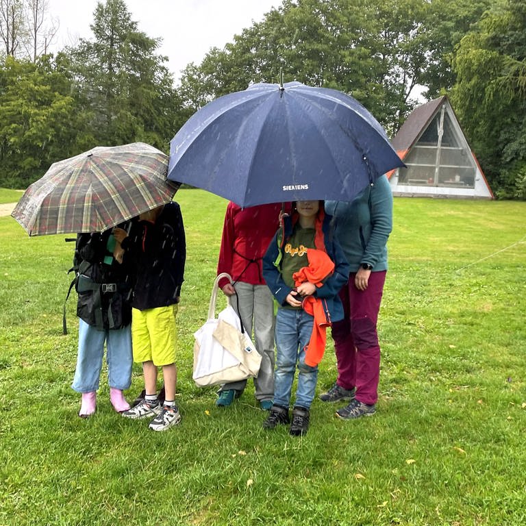 Fünf junge Camper auf einem Zeltplatz in Laichingen verstecken sich unter Schirmen, um dem Regen zu entkommen. (Foto: SWR, Catharina Straß)