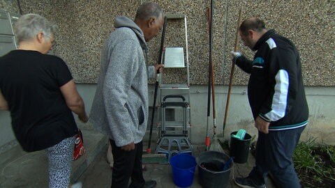 Menschen greifen nach an einer Hauswand stehenden Besen und Rechen. Rebellion mit Besenstil: Die Mieter und Mieterinnen in der Johannes-Palm-Straße 87 in Ulm greifen beherzt zu den "Waffen". Sie haben den Kampf um ihre Kehrwoche gewonnen, putzen jetzt weiter selbst und sparen sich dabei das Geld für einen externen Reinigungsservice.  (Foto: SWR, Frank Wiesner)