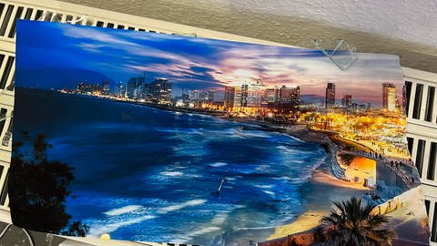 Plakat mit Ansicht einer abendlichen Stadt am Meer. Ein Plakat zeigt die Stadt Tel Aviv in Israel. Es wurde von einem Stein getroffen und in zwei Hälften gerissen. (Foto: SWR, Jannik Volz)