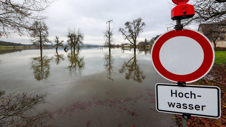 Hinter einem Durchfahrt verboten und Hochwasser-Hinweisschild, stehen Bäume im Wasser. Die Hochwasserlage bleibt in Teilen Baden-Württembergs weiterhin angespannt.