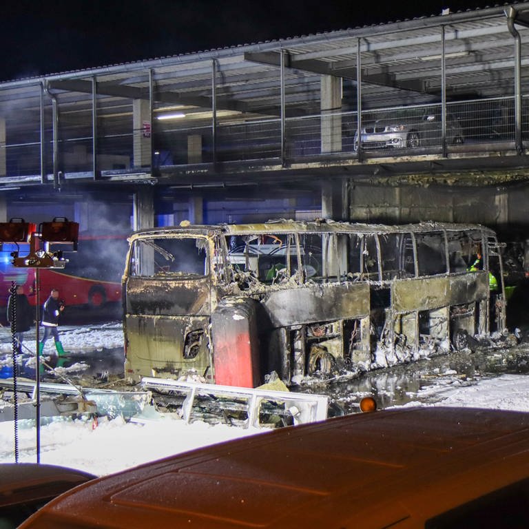 Ein komplett ausgebrannter Reisebus vor einer Halle. An der Seite stehen ein Feuerwehrauto und weitere geparkte Fahrzeuge. Bei dem Brand in Günzburg sind zwei Reisebusse komplett zerstört worden.