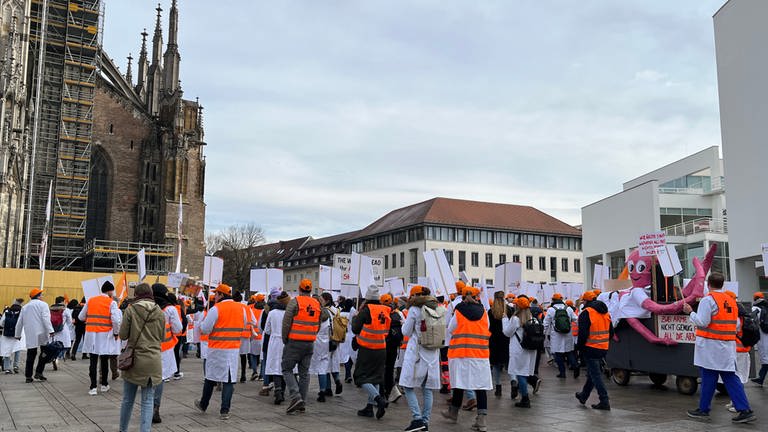 Ärztinnen und Ärzte der Uniklinik Ulm, teils in weißen Kitten, stehen mit Transparenten vor dem Ulmer Münster. Sie haben am Dienstagmittag für bessere Arbeitsbedingungen an der Uniklinik demonstriert.  (Foto: SWR)