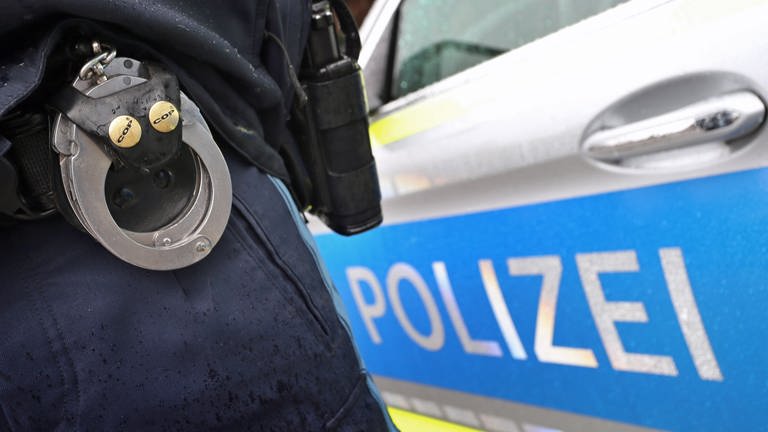 Ein Polizist zeigt Handschellen, im Hintergrund ein Polizeiwagen. Im Fall der in Günzburg gewaltsam zu Tode gekommenen Frau ist der 39-jährige Verdächtige in Untersuchungshaft. (Symbolbild) (Foto: dpa Bildfunk, picture alliance/dpa | Karl-Josef Hildenbrand)