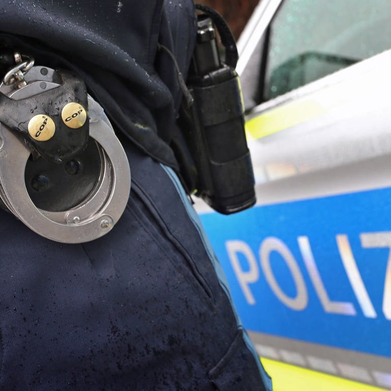 Ein Polizist zeigt Handschellen, im Hintergrund ein Polizeiwagen. Im Fall der in Günzburg gewaltsam zu Tode gekommenen Frau ist der 39-jährige Verdächtige in Untersuchungshaft. (Symbolbild)