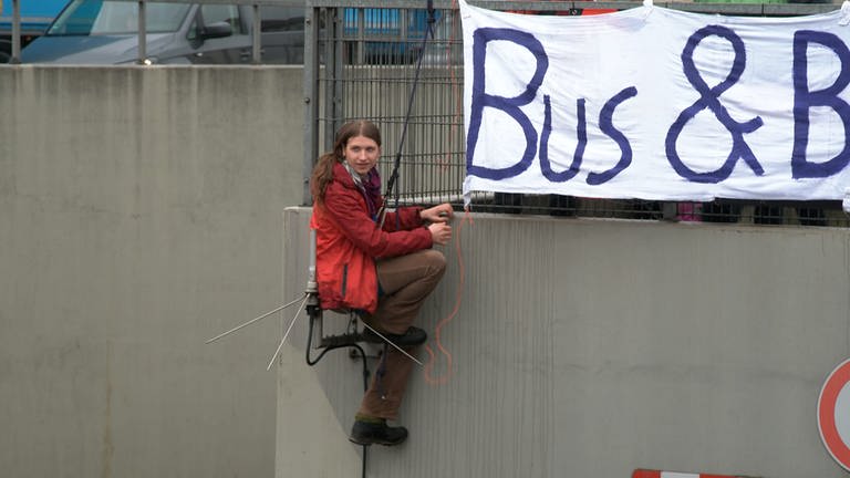 Samuel Bosch hängt in seinem Klettergurt an einer Unterführung. Über ihm ist ein Banner zu sehen.Auch vor Antritt seiner Haftstrafe lässt er sich nicht beirren. Heute hat er an der Unterführung B10B29 protestiert. (Foto: SWR)