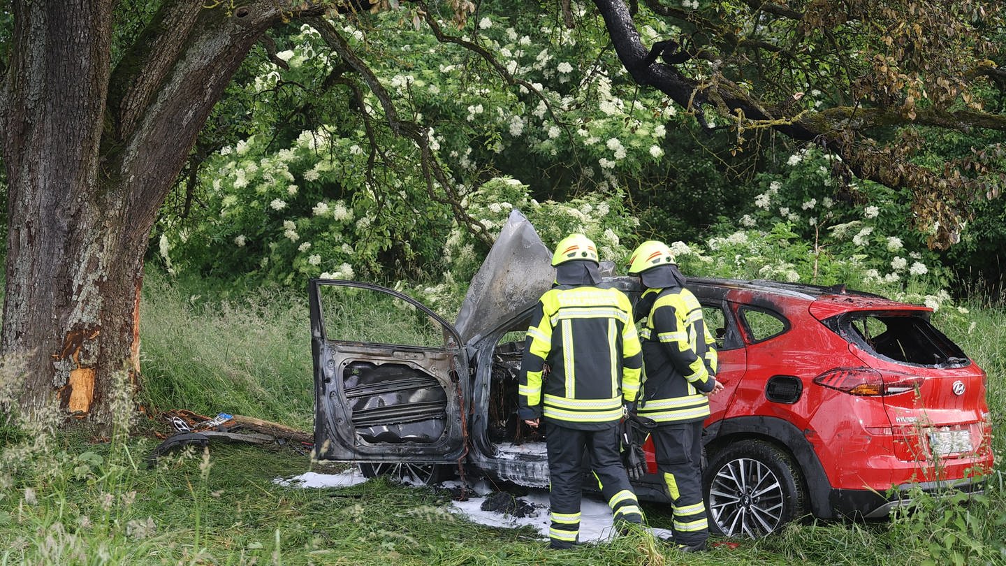 Zwei Feuerwehrleute stehen vor dem ausgebrannten Auto, dass am Donnerstag von der A8 gekommen und gegen einen Baum geprallt ist. (Foto: z-media)