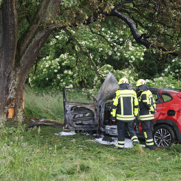 Zwei Feuerwehrleute stehen vor dem ausgebrannten Auto, dass am Donnerstag von der A8 gekommen und gegen einen Baum geprallt ist.