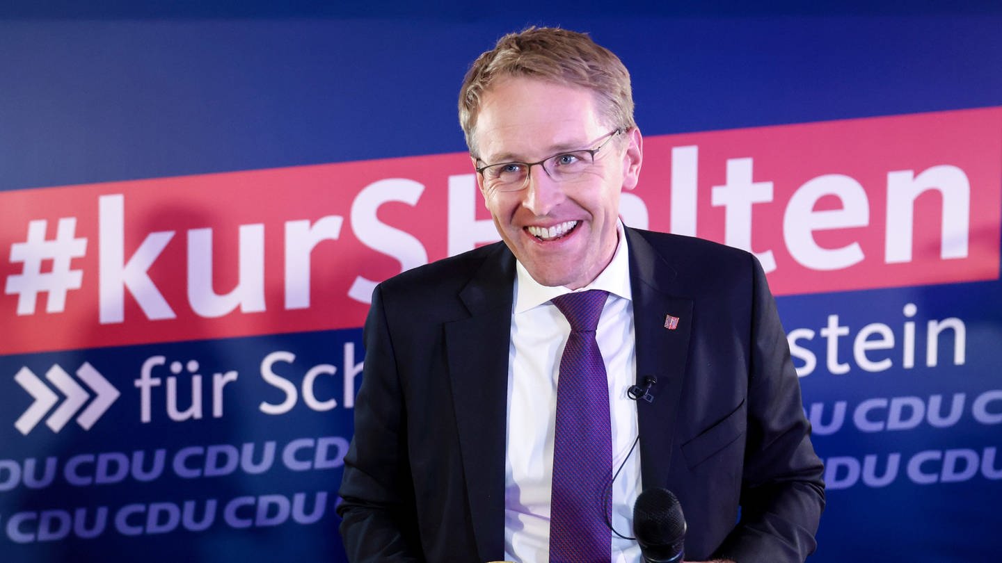 Daniel Günther (CDU) freut sich über den Sieg bei der Landtagswahl in Schleswig-Holstein und steht mit einem Bierglas in der Hand vor einem CDU-Wahlplakat.