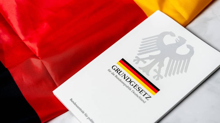 Ein Grundgesetz der Bundesrepublik Deutschland liegt auf einer deutschen Flagge in den Farben "schwarz-rot-gold". (Foto: IMAGO, IMAGO / Bihlmayerfotografie)