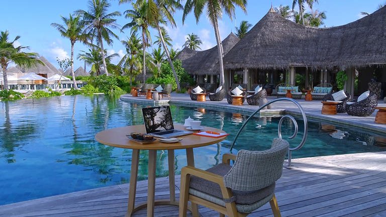 Ein Arbeitsplatz mit Laptop steht auf einem Café-Tisch mit Sessel neben einem Pool, umringt mit Palmen und Hütten einer Hotel-Anlage auf den Malediven. (Foto: dpa Bildfunk, picture alliance/dpa/The Nautilus Maldives | -)