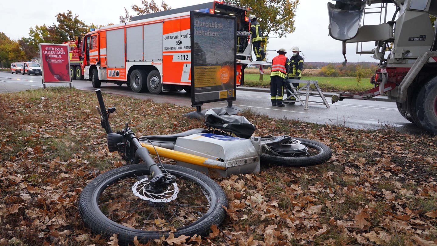 Ein E-Bike liegt nach einem Unfall hinter einer Bushaltestelle auf einer Wiese. Im Hintergrund sieht man ein Feuerwehrauto und Feuerwehrmänner. (Foto: dpa Bildfunk, picture alliance/dpa | Andreas Rosar)