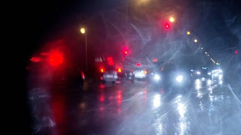 Durch eine Frontscheibe ist bei Regen eine unübersichtliche Verkehrssituation im Dunkeln zu sehen.