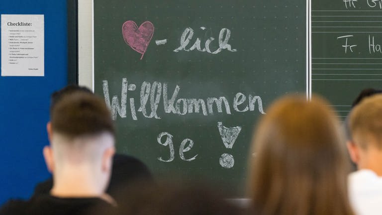 "Herzlich Willkommen 9e" Schriftzug an der Tafel eines Klassenzimmers begrüßt die Schüler nach den Ferien