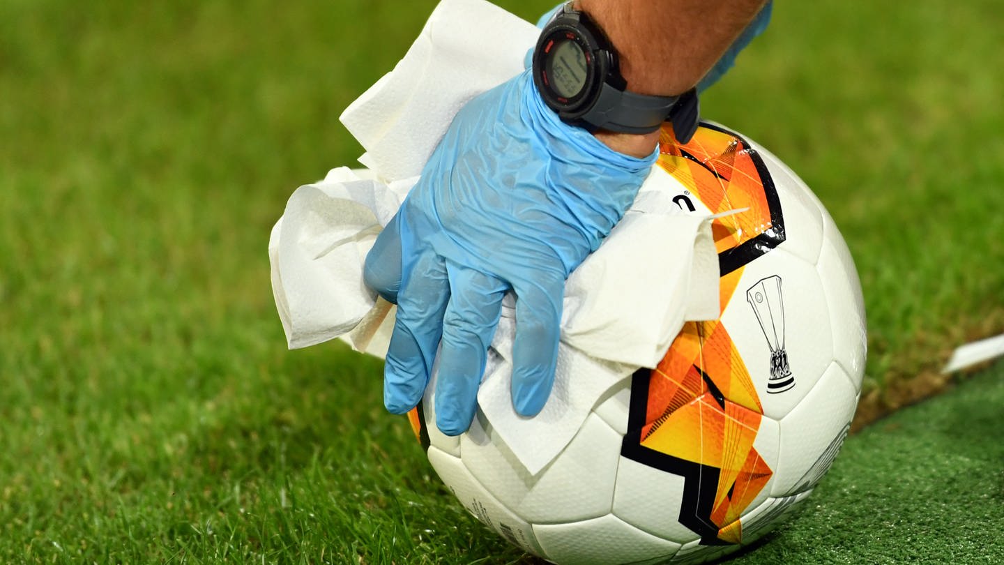 Ein Helfer mit blauen Latexhandschuhen desinfiziert einen Fußball (Foto: dpa Bildfunk, picture alliance/ dpa/ Marius Becker)