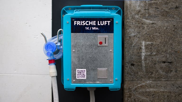 Ein Automat mit Aufschrift "Frische Luft" für einen Euro pro Minute und einer Atemmaske hängt an einem Haus. Ein QR-Code am Automaten führt zum Eintrag "Luftverschmutzung" in der Online-Enzyklopädie Wikipedia.