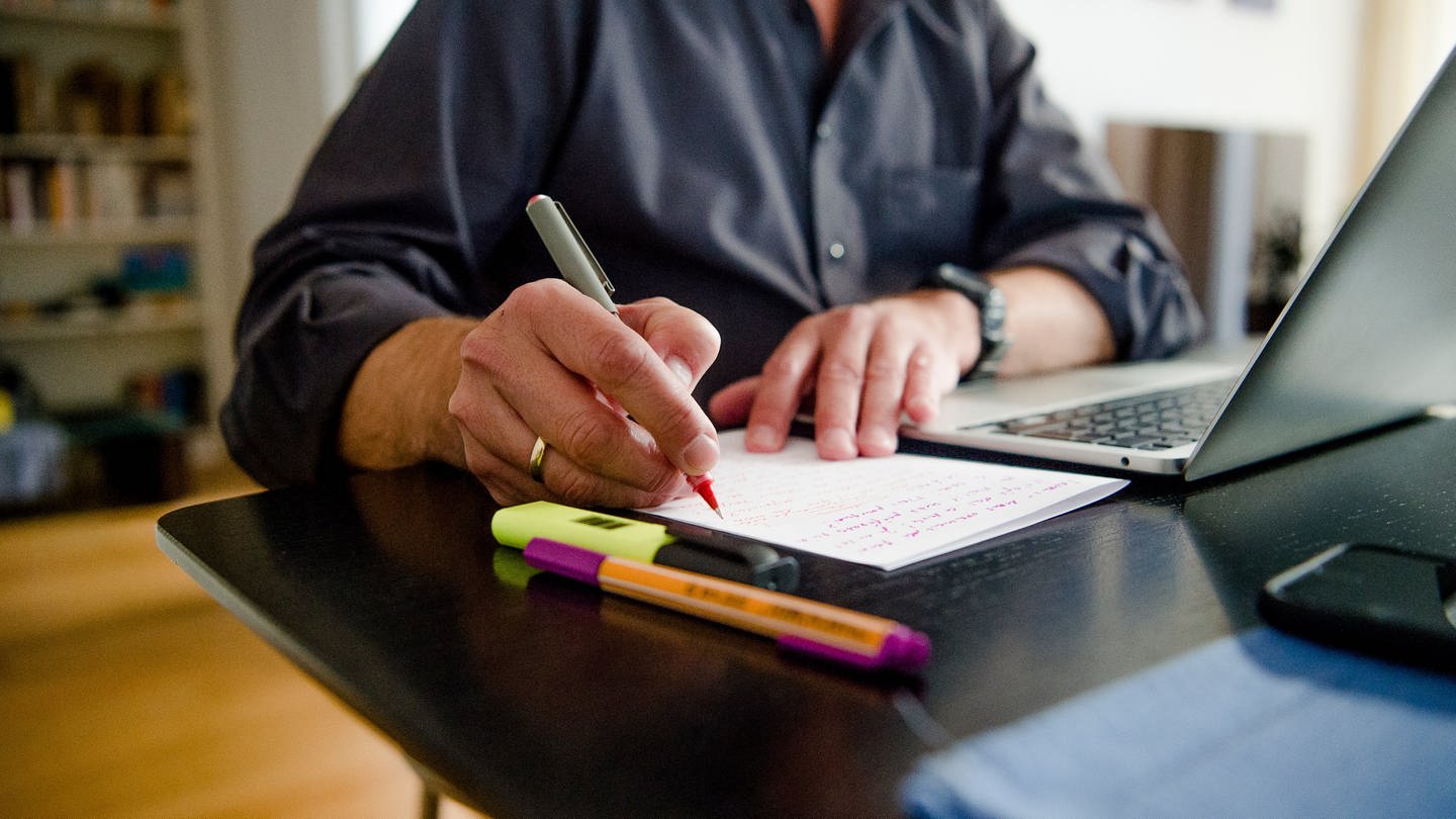 Ein Mann sitzt zuhause an einem Esstisch. Er schreibt mit einem Stift auf ein Blatt Papier neben einem Laptop. (Foto: dpa Bildfunk, picture alliance/dpa | Finn Winkler)