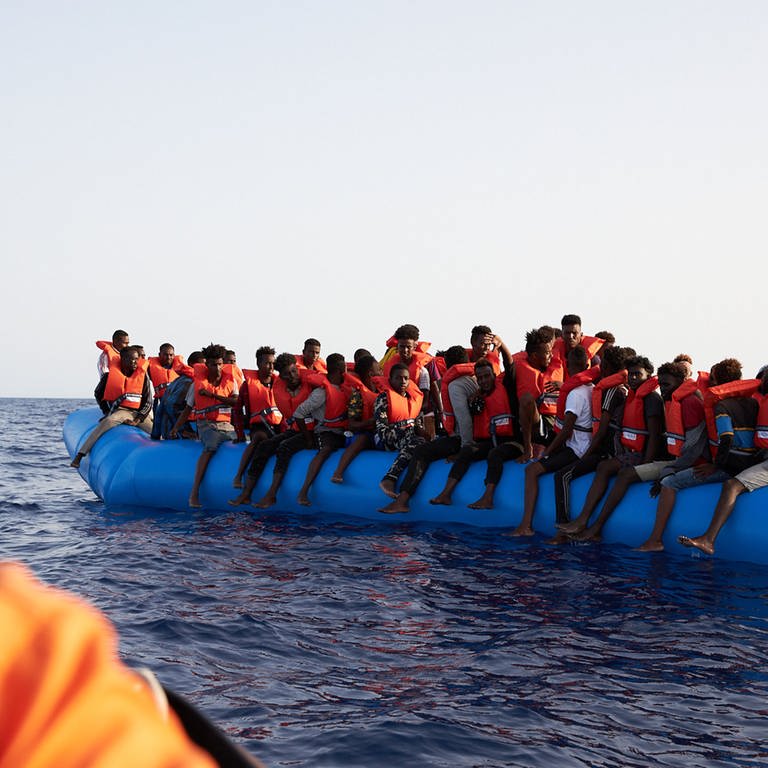 Ein Seenotretter in Schutzkleidung schaut zu einem vollen Flüchtlingsboot.