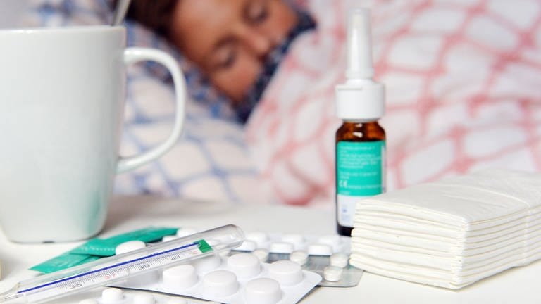Medikamente und ein Fieberthermometer liegen auf einem Nachttisch - im Hintergrund liegt eine Frau mit Erkältung im Bett und schläft.
