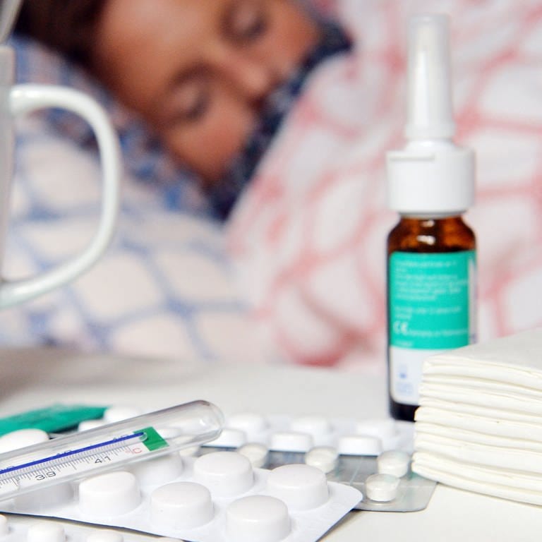 Medikamente und ein Fieberthermometer liegen auf einem Nachttisch - im Hintergrund liegt eine Frau mit Erkältung im Bett und schläft. (Foto: dpa Bildfunk, picture alliance/dpa | Maurizio Gambarini)