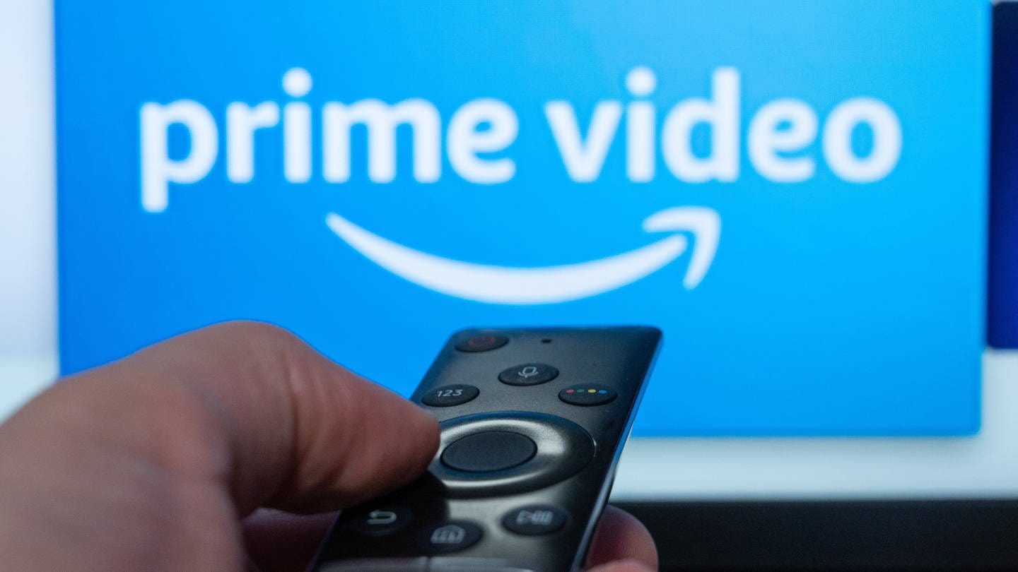 Ein Mensch hält eine Fernbedienung und richtet sie auf einen Bildschirm, auf dem das Amazon Prime Video Logo zu sehen ist (Foto: dpa Bildfunk, picture alliance/dpa | Daniel Reinhardt)