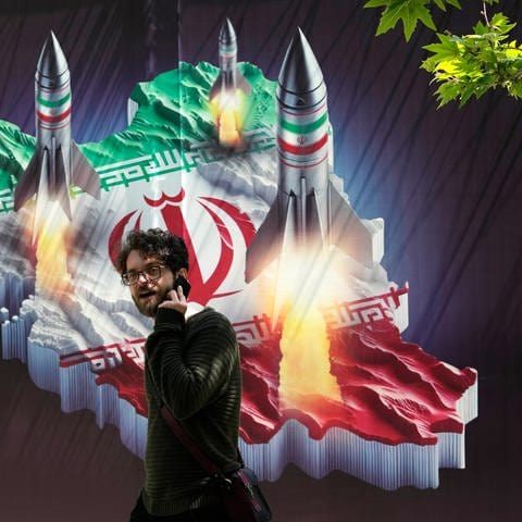 Ein Passant, der mit einem Handy telefoniert, geht an einem Banner vorbei, das den Abschuss von Raketen von der iranischen Landkarte im Norden Teherans zeigt. (Foto: dpa Bildfunk, picture alliance/dpa/AP | Vahid Salemi)