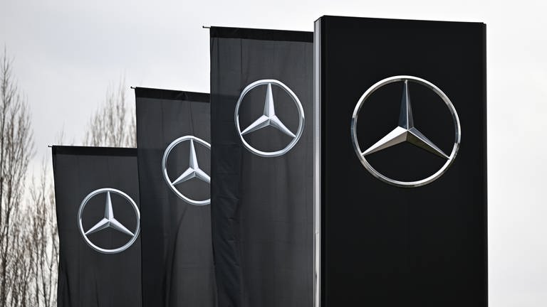 Das Logo der Automarke Mercedes-Benz weht an Fahnen vor einer Mercedes-Benz Niederlassung. 