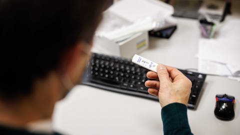 Ein Mitarbeiter an einem Schreibtisch schaut auf das Testergebnis für einen Corona-Schnelltest eines anderen Mitarbeiters. (Foto: dpa Bildfunk, picture alliance/dpa | Matthias Balk)