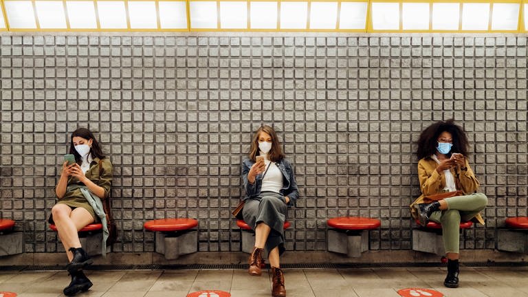 Drei Frauen sitzen an einer U-Bahnstation mit ihren Handys auf einer Bank. Handys sind für die meisten aus dem Alltag nicht mehr wegzudenken: Doch sie rücken zunehmend ins Visir von Hackern. (Foto: IMAGO, IMAGO / Westend61)