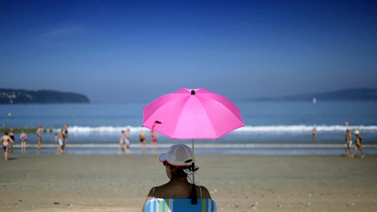 Eine Frau sitzt unter einem pinken Sonnenschirm am Strand.