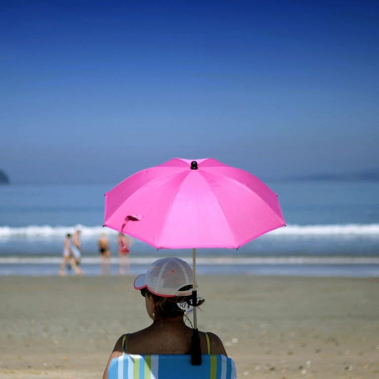 Eine Frau sitzt unter einem pinken Sonnenschirm am Strand. (Foto: SWR,  )