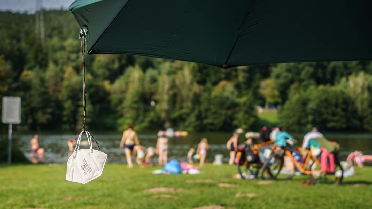 Eine FFP2-Maske baumelt von einem Sonnenschirm. Im Hintergrund tummeln sich bei sommerlichem Wetter die Menschen zum Baden und Surfen.