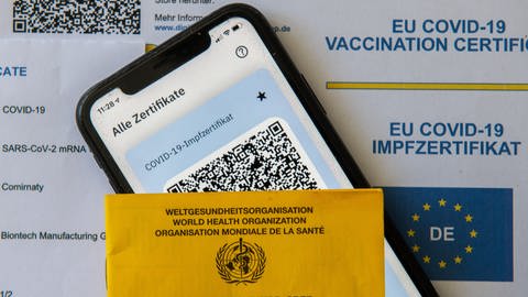 Ein Impfpass und ein Smartphone, auf dem die App CovPass läuft, liegen auf einem Impfzertifikat. (Foto: picture-alliance / Reportdienste, Picture Alliance)